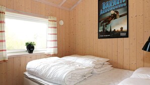 A room with wooden walls, a bed with white bedding, a window with curtains, a poster on the wall, and a small plant on the windowsill.