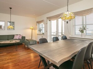 A modern living room with a dining table, sofa, and chairs, wooden flooring, and large windows with curtains.