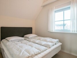 Un dormitorio con cama, ropa de cama blanca, cabecera oscura y ventana con persianas.