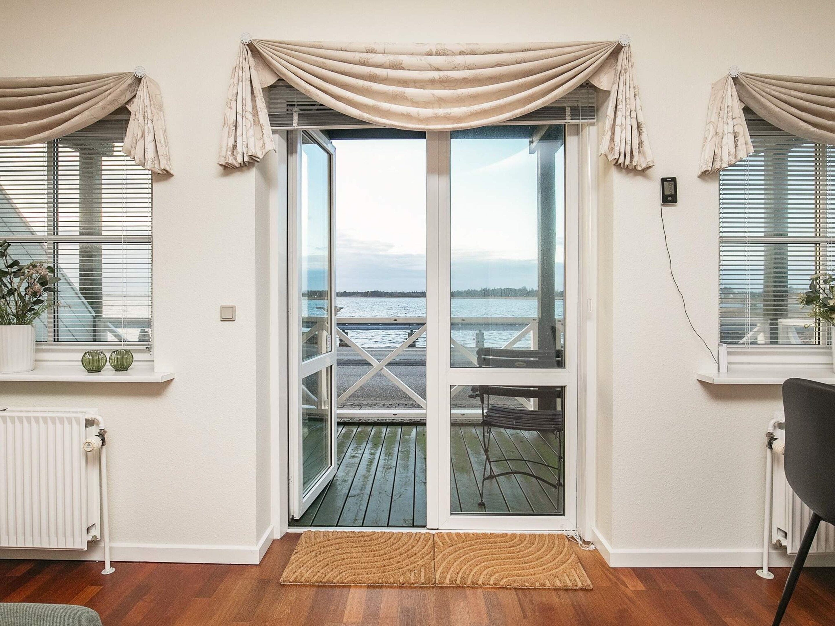 A room with a sliding glass door leading to a balcony with a view of a body of water. There are two windows with curtains, a radiator, and a doormat.