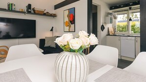 A modern living room with a white dining table, a striped vase with white roses, a flat-screen TV, and a kitchen visible through an open door. - 4 Star Holiday Home in Vaeggerlose (Vaeggerlose)
