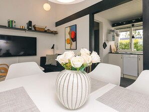 Ein modernes Wohnzimmer mit einem weißen Esstisch, einer gestreiften Vase mit weißen Rosen, einem Flachbildfernseher und einer durchgehenden Tür erkennbaren Küche.