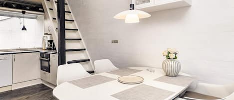 A modern kitchen with a dining area, featuring a white table with placemats, a vase with flowers, and a staircase leading to an upper level.