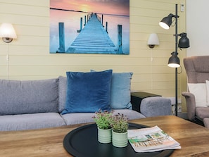 A room with a sofa, a coffee table with magazines and plants, a painting of a pier, and wall-mounted lamps.