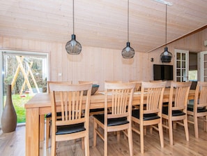 A wooden dining area with a long table and chairs, overhead pendant lights, and a view of the outdoors.