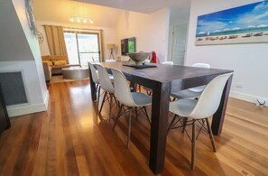 A dining area with a dark wooden table and white chairs, a sofa, and a TV in the background.
