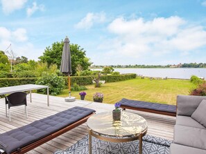 An outdoor deck with a glass-top table, cushioned seating, and a view of a lake. - 5 Star Holiday Home in Praesto (Praesto)