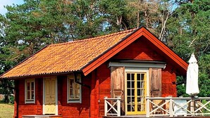 A small, red wooden cabin with a porch and a yellow door, surrounded by trees. - 6 Person Holiday Home in Soderakra (Soderakra)