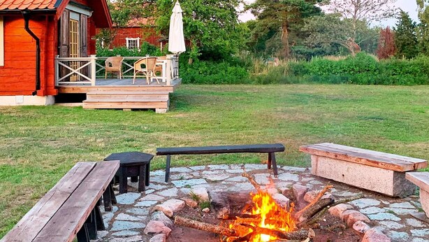 A fire pit surrounded by stone tiles, wooden benches, and a patio area with a red building in the background. - 6 Person Holiday Home in Soderakra (Soderakra)
