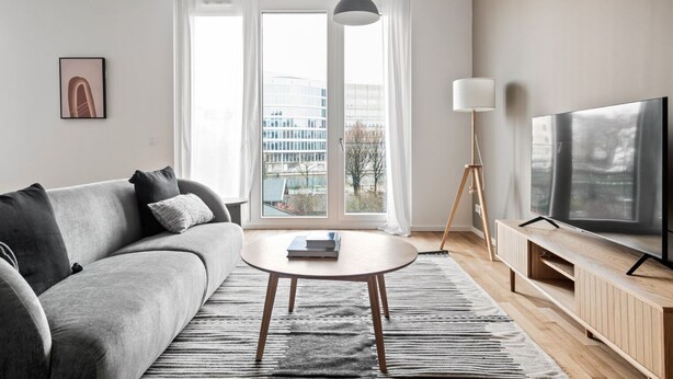 Un salon moderne avec un canapé gris, une table basse ronde, un téléviseur posé sur un meuble en bois et une lampe sur pied.