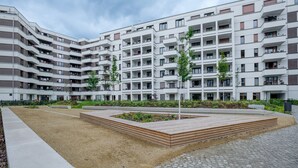 Un immeuble d’appartements moderne doté de nombreux balcons, d’une cour aménagée avec des jardinières en bois et d’un chemin piétonnier pavé.