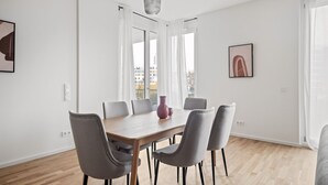 Une salle à manger moderne avec une table en bois et des chaises grises, de grandes fenêtres avec des rideaux blancs et une vue sur la ville.
