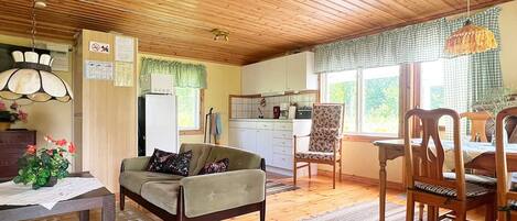 A cozy living area with a sofa, a dining table, and chairs, a kitchen with white cabinets, and a window with a checkered curtain.