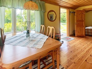 Ein Holzespeisetisch mit Stühlen, einer Vase und einer Tischmatte. Ein Fenster mit Vorhängen, eine zur Außenwelt führende Tür und ein Spiegel an der Wand sind zu sehen.