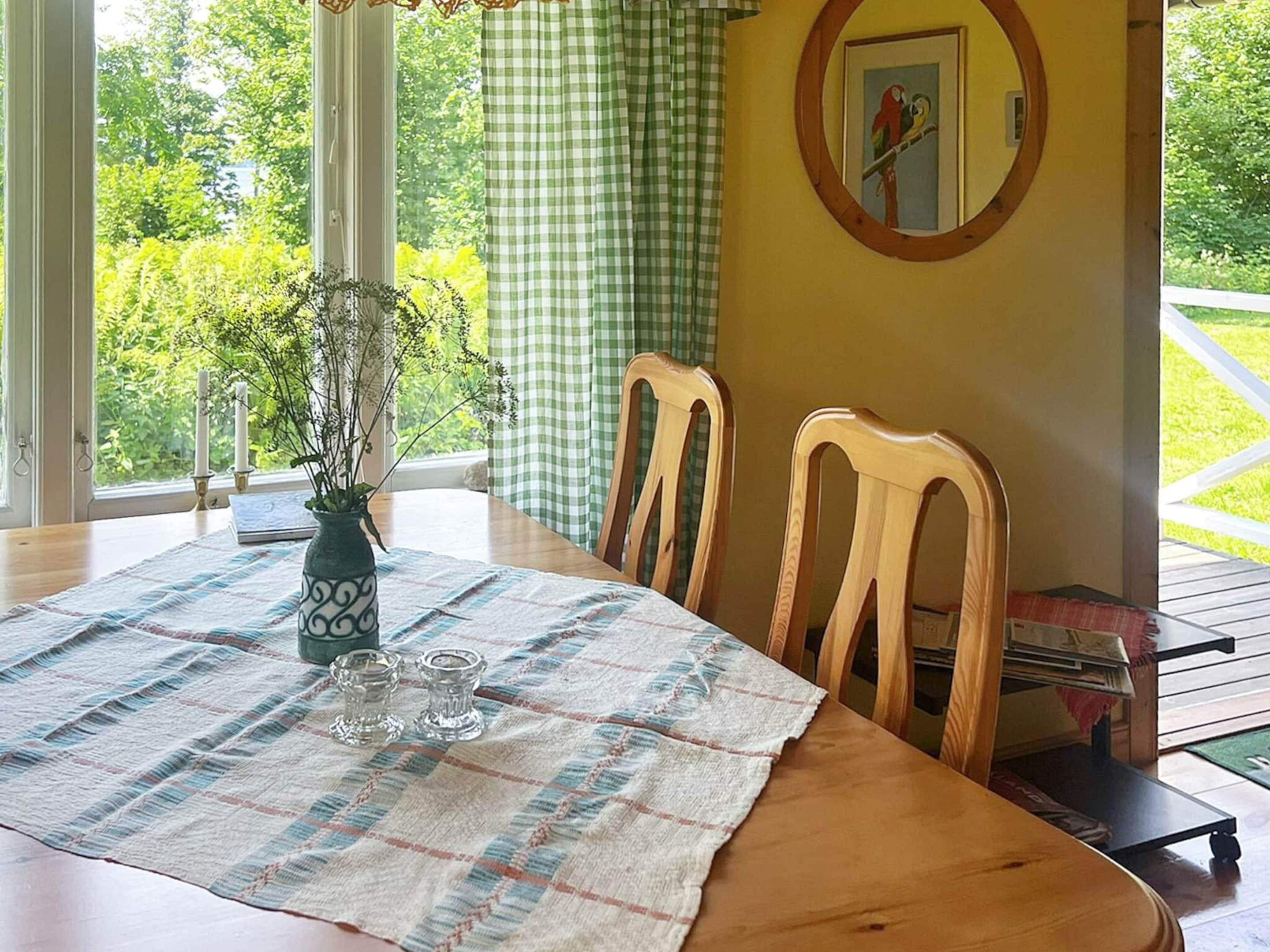 A dining area with a table set for two, a vase with flowers, and a view of greenery outside.