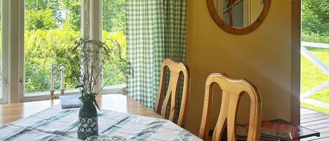 A dining area with a table set for two, a vase with flowers, and a view of greenery outside.