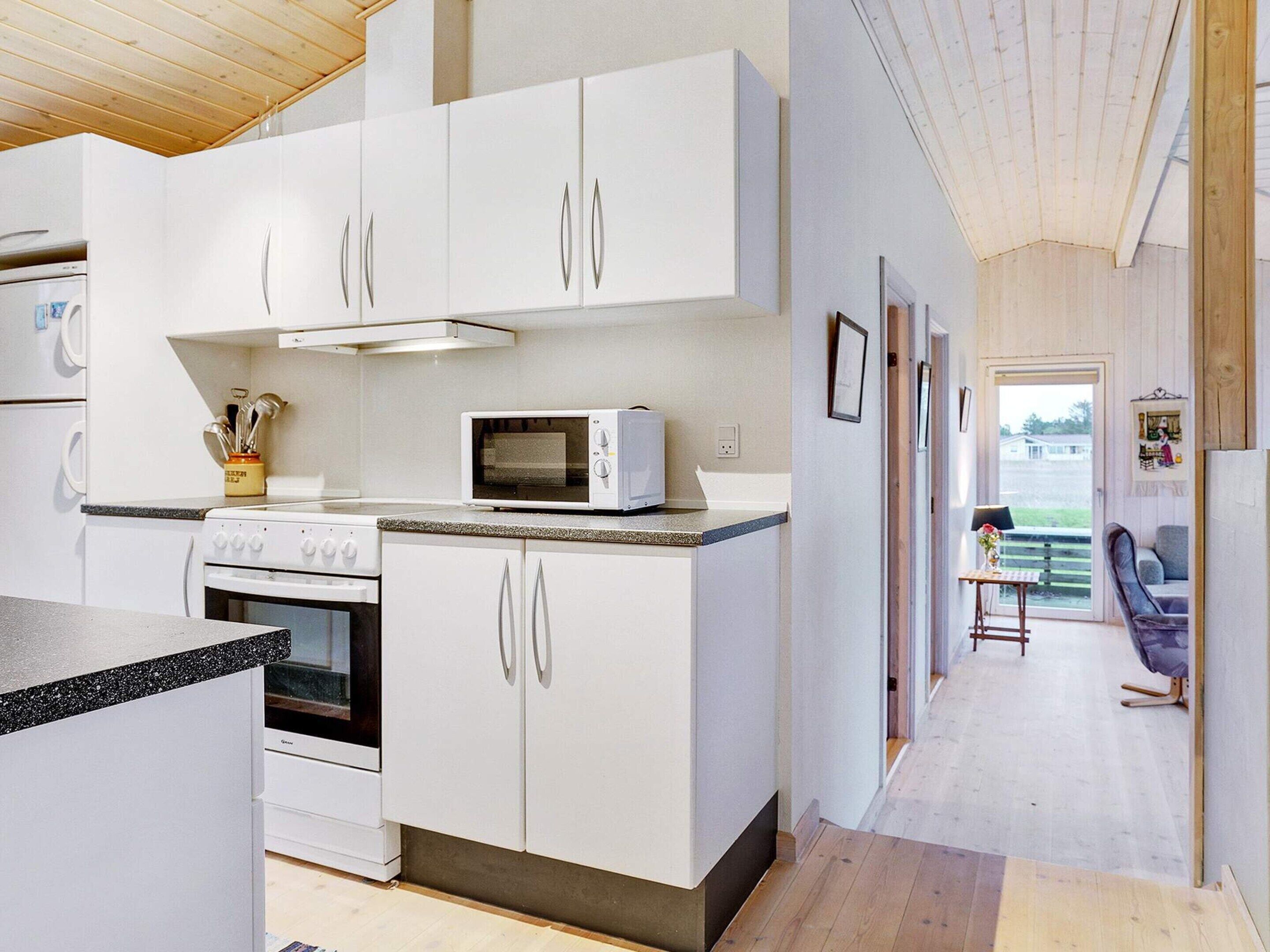 A modern kitchen with white cabinets, a microwave, and an oven. There is a wooden ceiling and a view of a living area with a sofa and a table.