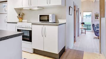 A modern kitchen with white cabinets, a microwave, and an oven. There is a wooden ceiling and a view of a living area with a sofa and a table.