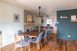 A modern dining area with a wooden table and grey upholstered chairs, a chandelier, and a kitchen in the background. - Denver Escape-10: Patio, Hot Tub, JamRoom, FirePit (Denver)
