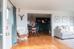 A modern living room with a grey sofa, a dining area with a wooden table and chairs, and a kitchen in the background. - Denver Escape-10: Patio, Hot Tub, JamRoom, FirePit (Denver)