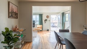 A modern living room with a wooden dining table, a sofa, and a coffee table. There is a plant on the left and a bookshelf with books and a small plant.