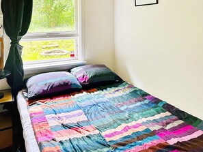 Una cama con colcha colorida, una ventana con cortinas y vistas al exterior con vegetación.
