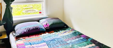 A bed with a colorful quilt, a window with curtains, and a view of greenery outside.