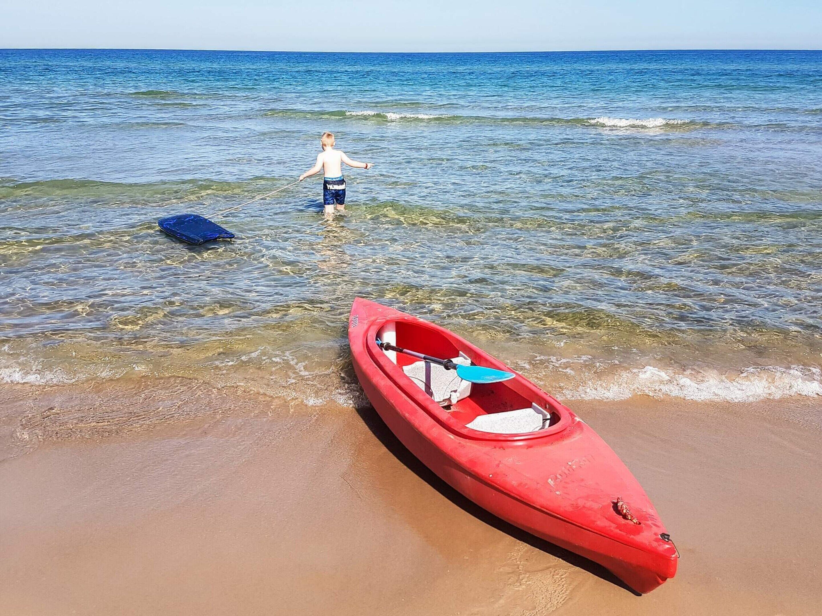 Un kayak rouge sur une plage de sable, avec une eau claire et une personne à l’arrière-plan.