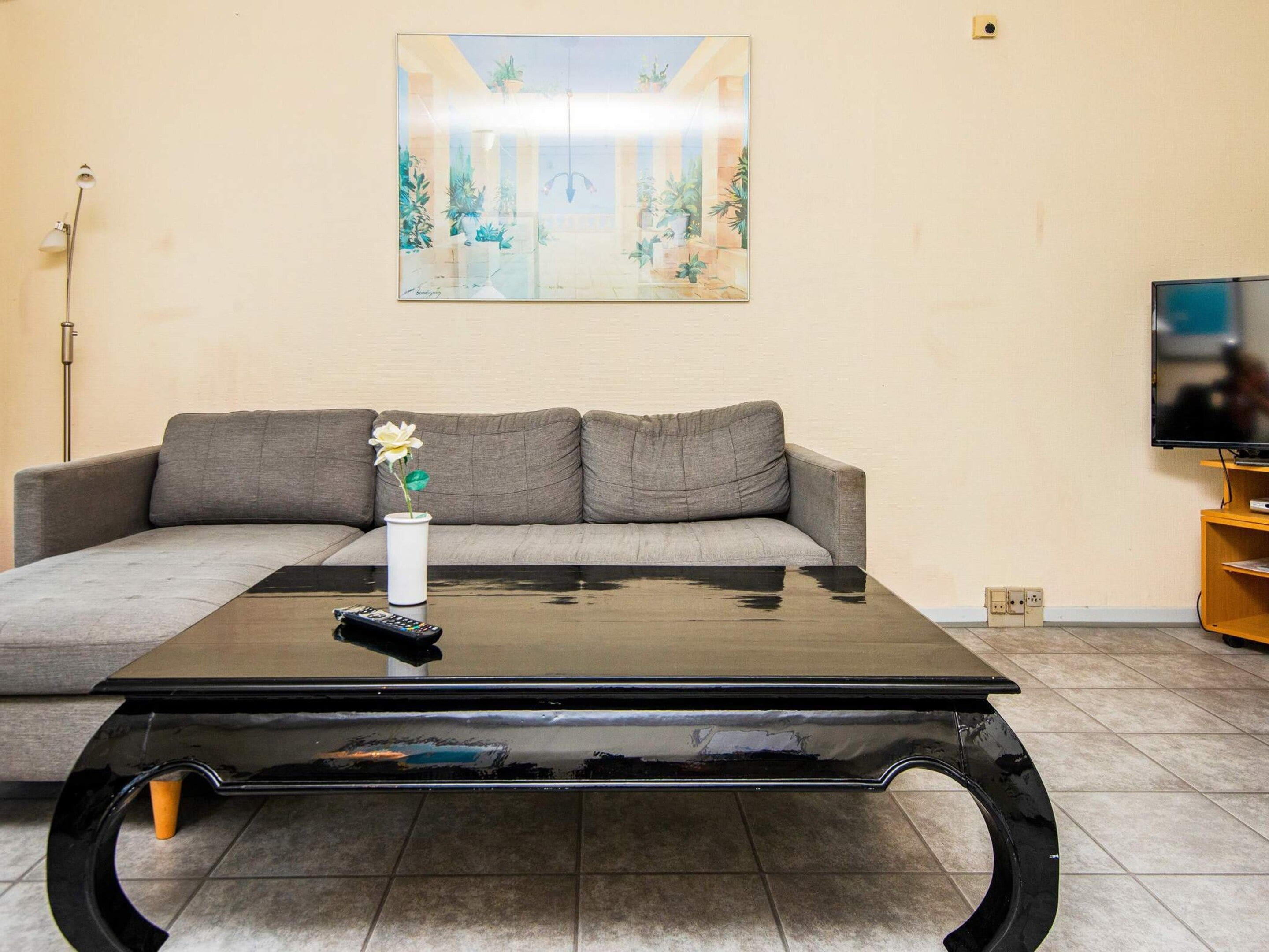 A living room with a grey sofa, a coffee table, a vase with a flower, a remote control, and a painting on the wall.