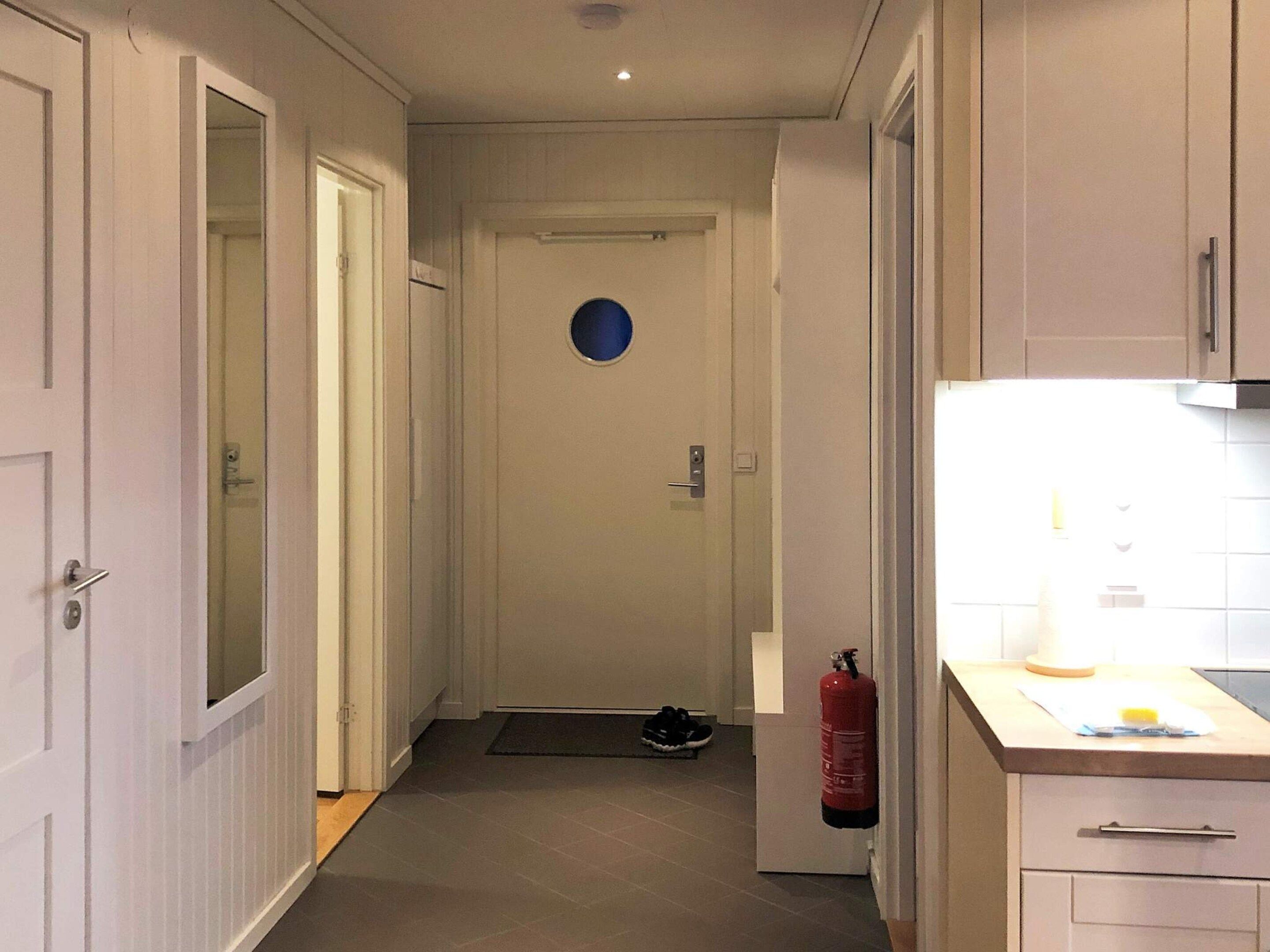 Un couloir étroit mène à une porte percée d’une ouverture circulaire. Un extincteur est fixé au mur, et un petit tapis recouvre le sol.