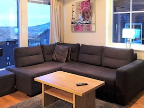 A modern living room with a sectional sofa, a wooden coffee table, and a view of a cityscape through large windows. - 4 Star Holiday Home in Salen (Salen)