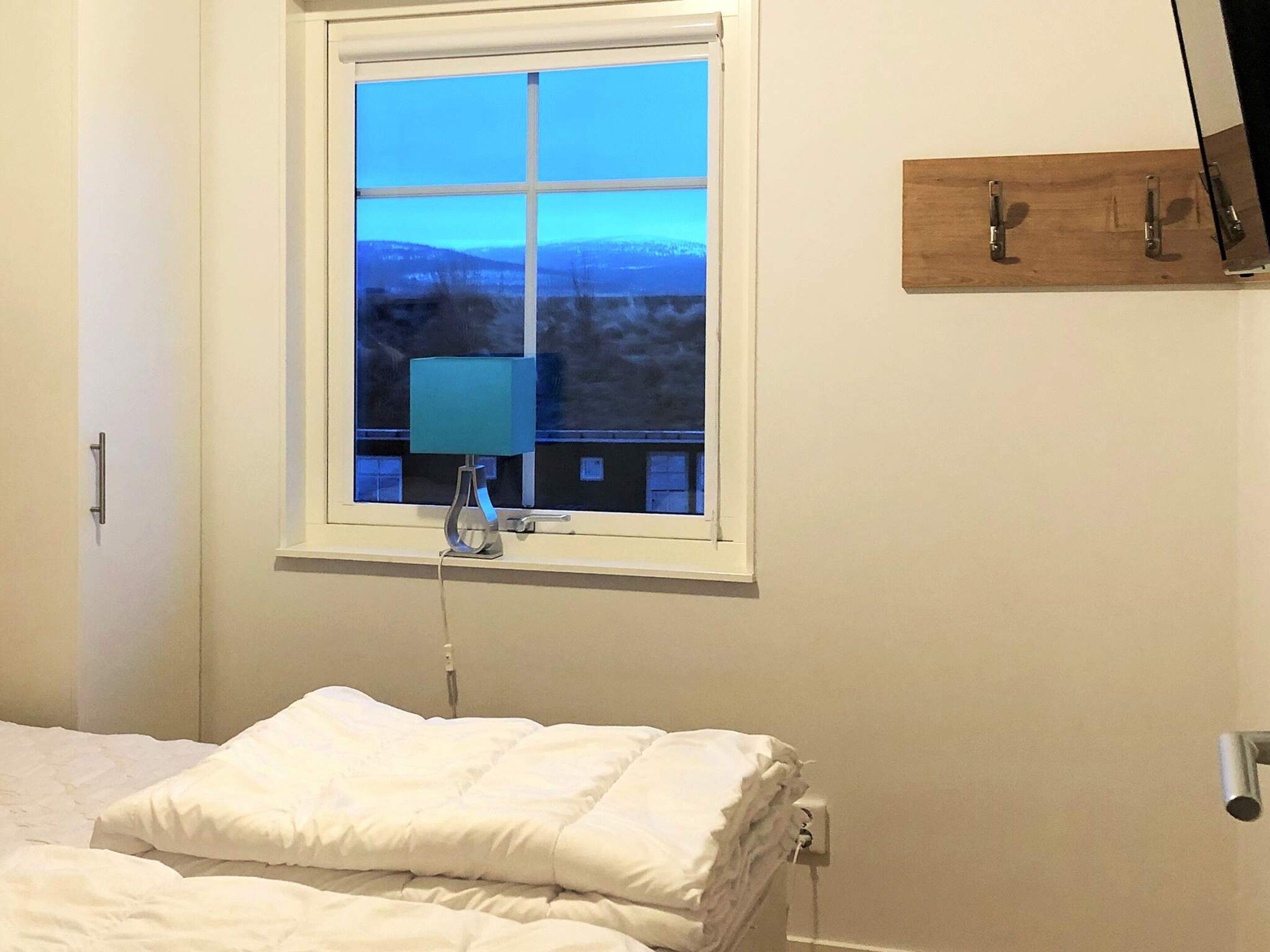 A bedroom with a bed, a window with a view, a bedside table, and a wall-mounted shelf.
