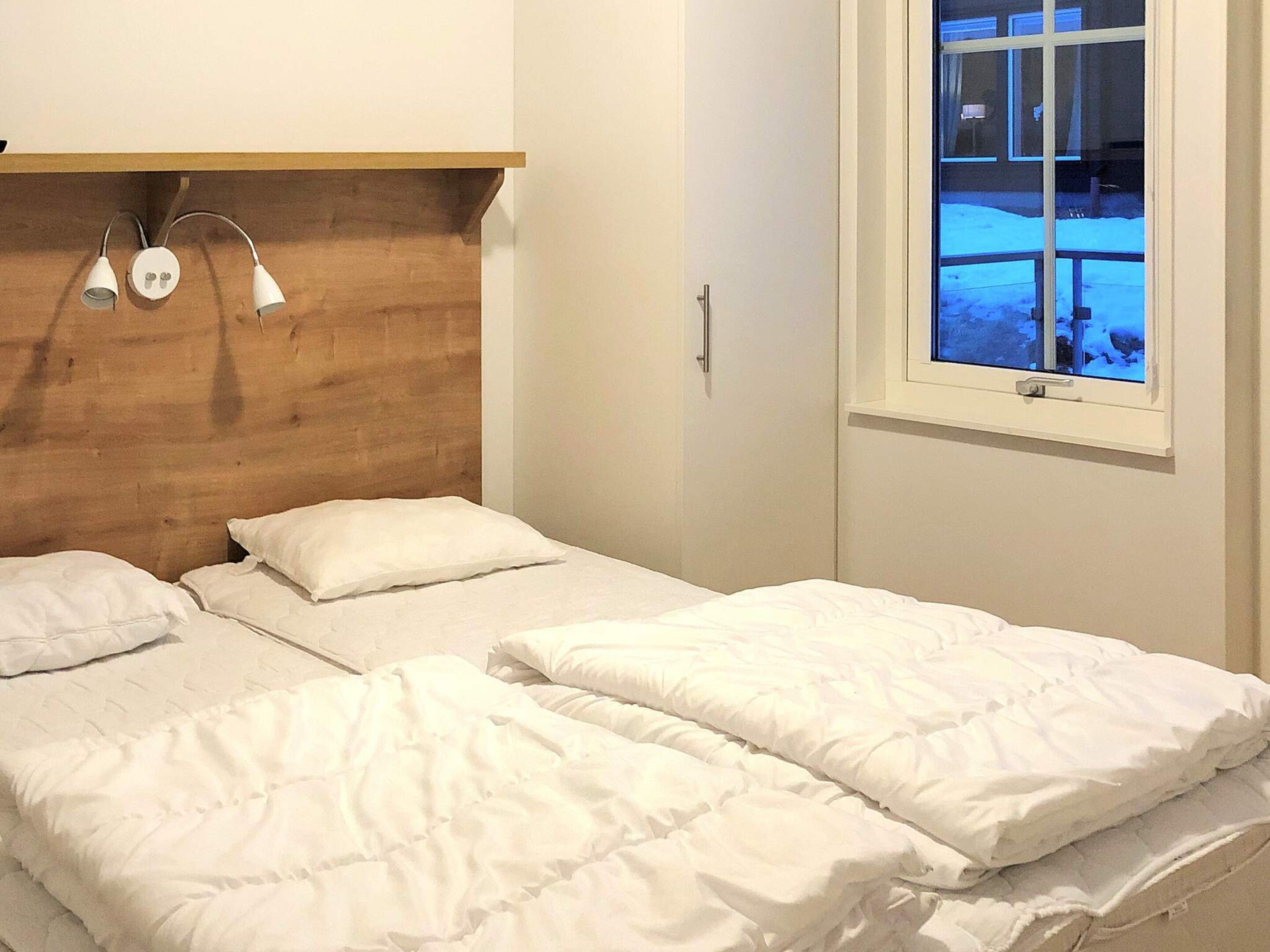 Une chambre à coucher avec une tête de lit en bois, deux lits avec du linge de lit blanc, une table de chevet et une fenêtre donnant sur un paysage enneigé.