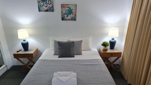 Un llit ben fet amb un llençol gris, dues taules de nit amb làmpades, una petita planta en maceta i una obra d'art a la paret.