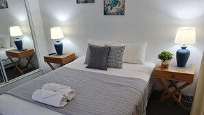 Un llit ben fet amb un llençol gris, dues taules de nit amb làmpades, una petita planta en test i un mirall a la paret.