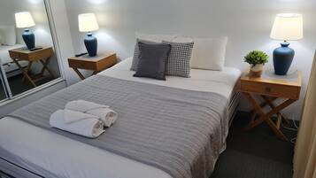 A neatly made bed with a grey quilt, two bedside tables with lamps, a small potted plant, and a mirror on the wall.