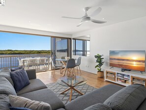 Ein modernes Wohnzimmer mit Balkon und Blick auf ein Gewässer, inklusive Deckenventilator und Fernseher auf einer Holzkassette.