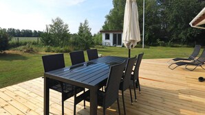 A wooden deck with a table and chairs, an umbrella, and a view of a house and trees in the background. - 8 Person Holiday Home in Laeso (Læsø)