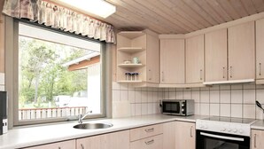 A kitchen with wooden cabinets, a microwave, an oven, a sink, and a window with a view of trees and a building outside. - 8 Person Holiday Home in Laeso (Læsø)