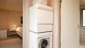 A compact laundry room with a stacked washer and dryer, a small bedside table, and a window with a view. - 8 Person Holiday Home in Laeso (Læsø)