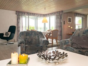 Un salon avec un plafond en bois, deux fauteuils, un canapé, une table à manger avec des chaises et une fenêtre avec des rideaux.