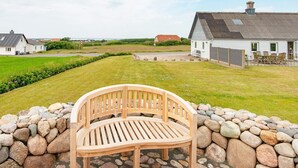 Een houten bank in een tuin met een stenen muur, een wit huis met een grijs dak en een heldere lucht.