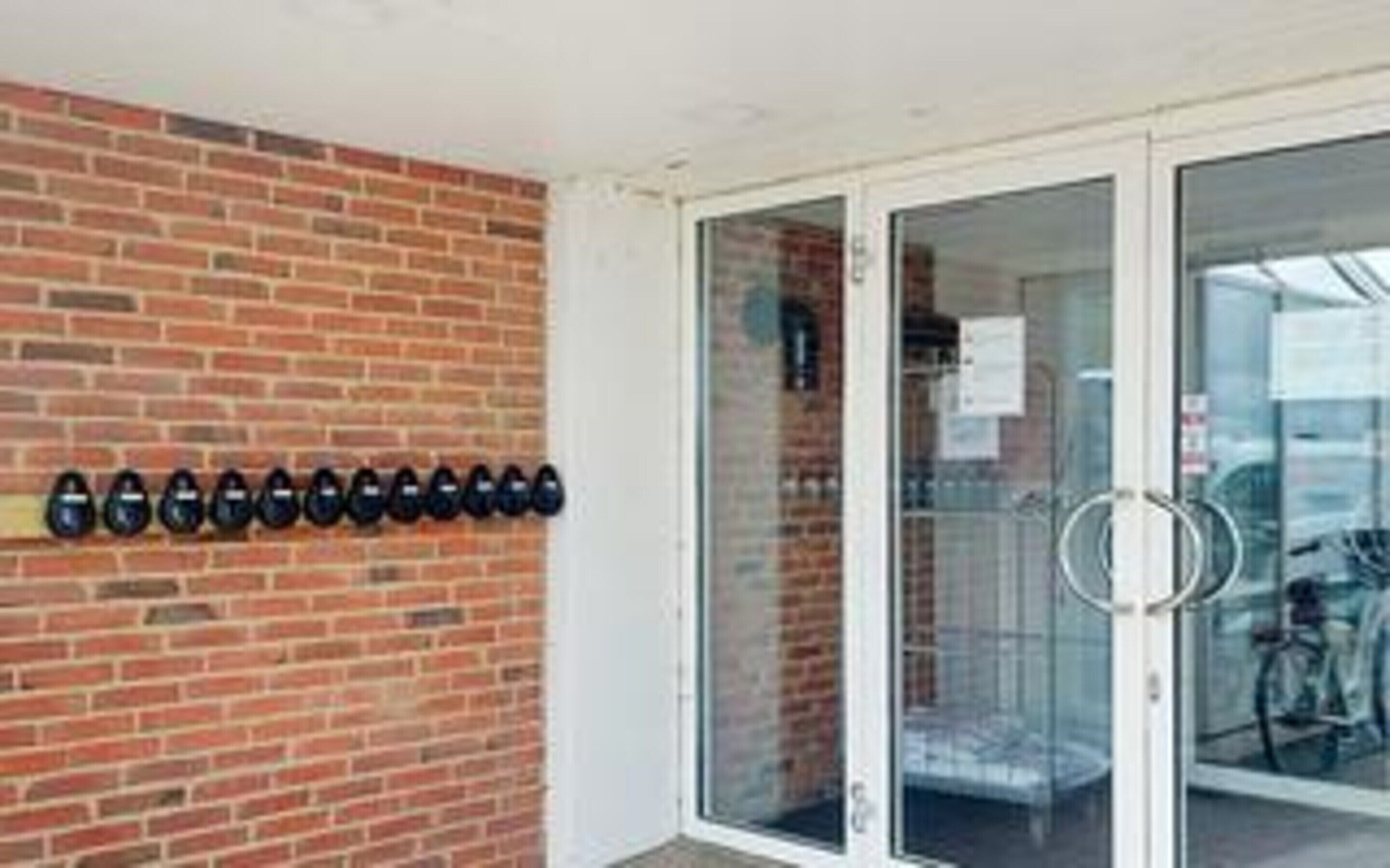 A brick wall with a row of black objects, a glass door with a handle, and a glimpse of a bicycle inside.