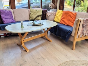 Et hyggeligt opholdsområde med et rundt bord, en sofa med farverige puder og udsigt gennem store vinduer.