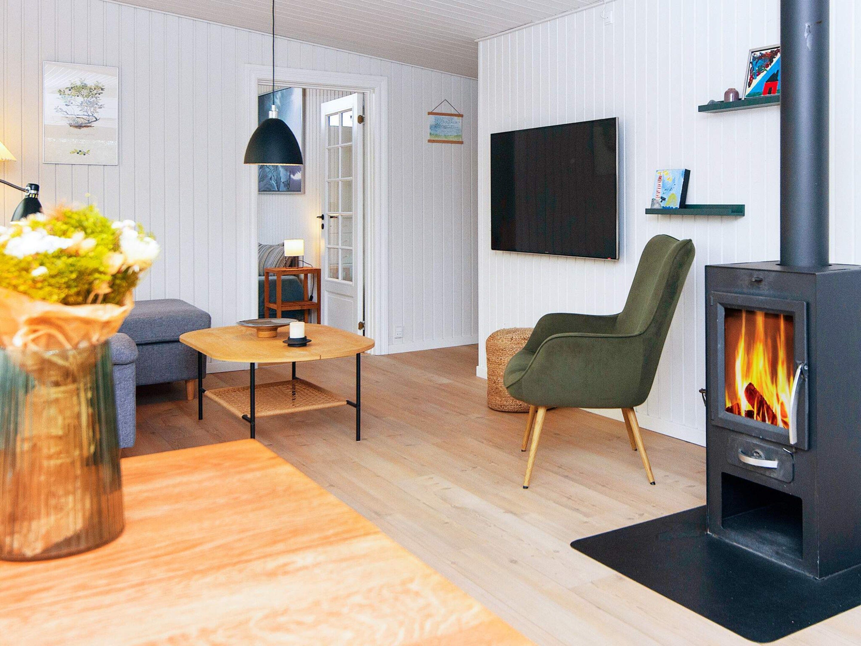 Een moderne woonkamer met een bank, een ronde tafel, een groene fauteuil, een televisie en een houtkachel.