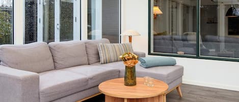 Un salon moderne avec un canapé gris, une table basse ronde en bois et une grande fenêtre donnant sur une autre pièce.