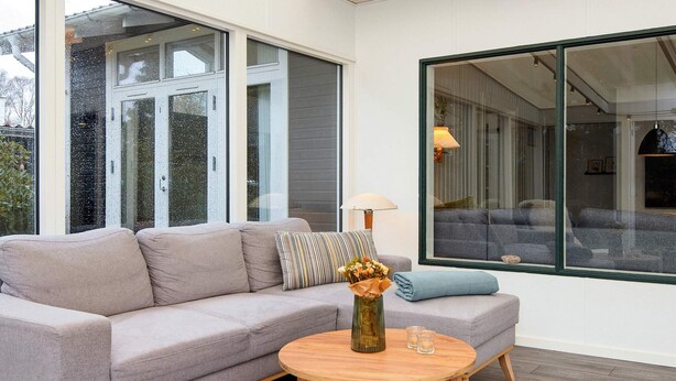 Uma sala de estar moderna com um sofá cinzento, uma mesa de centro redonda de madeira e uma janela grande com vista para outro quarto.