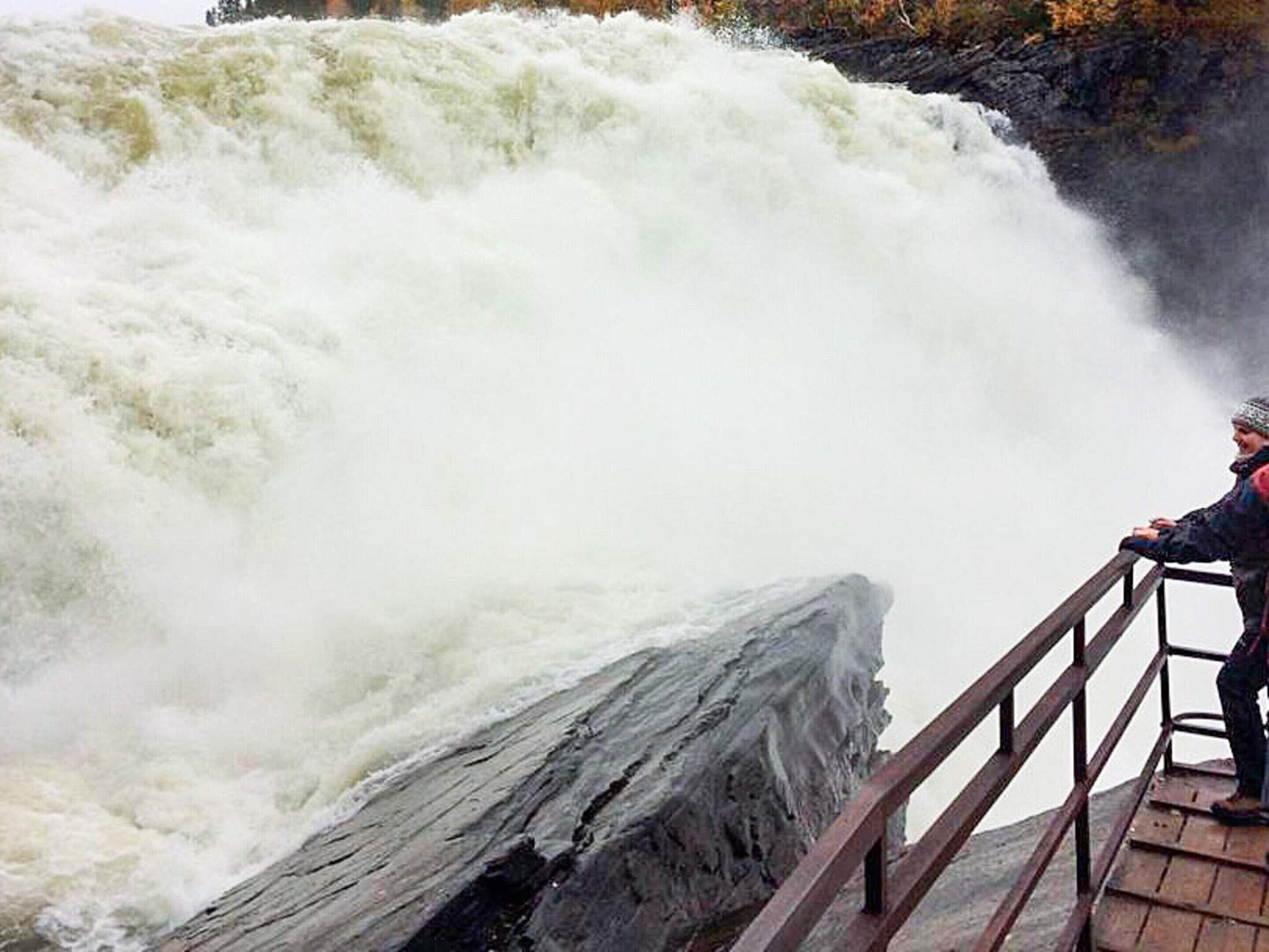 A person standing on a wooden platform with a railing, overlooking a large waterfall.