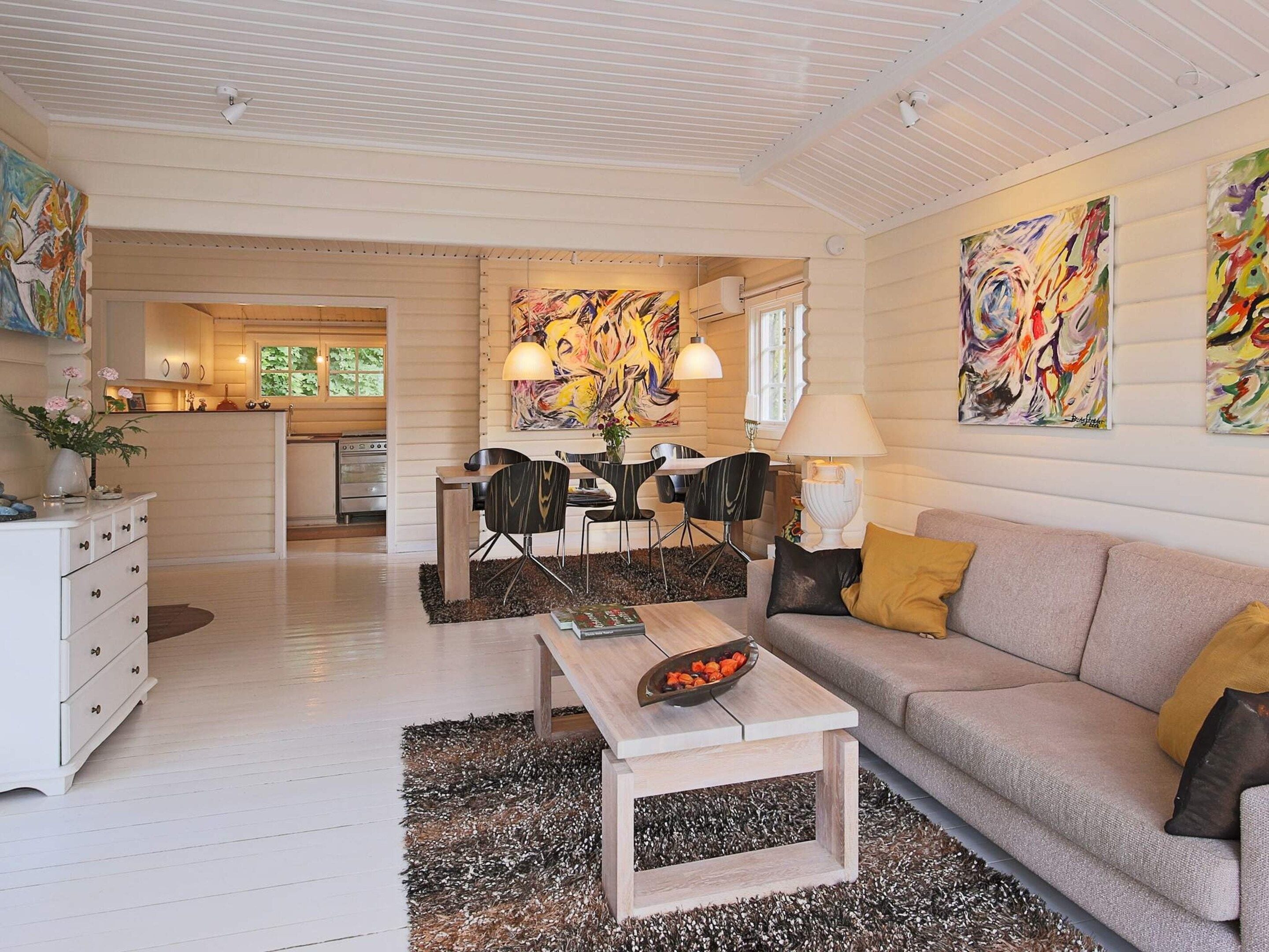 A spacious living area with a sofa, coffee table, and dining table. There is a kitchen in the background with a window and a painting on the wall.