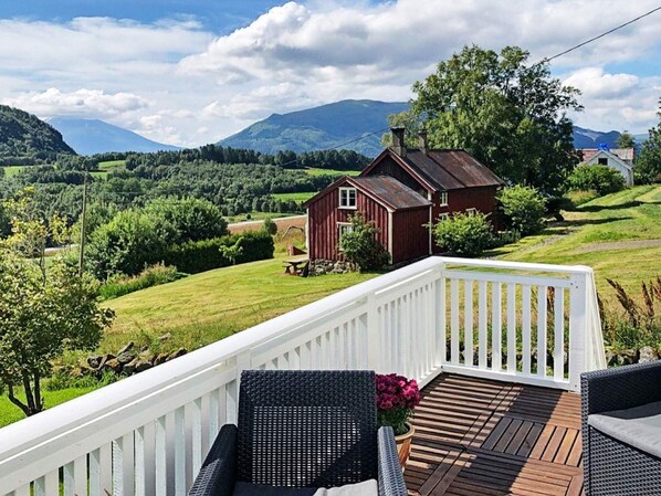 A wooden deck with wicker chairs, a red barn, and a scenic view of green hills and mountains. - 6 Person Holiday Home in Averoy (Averoy)